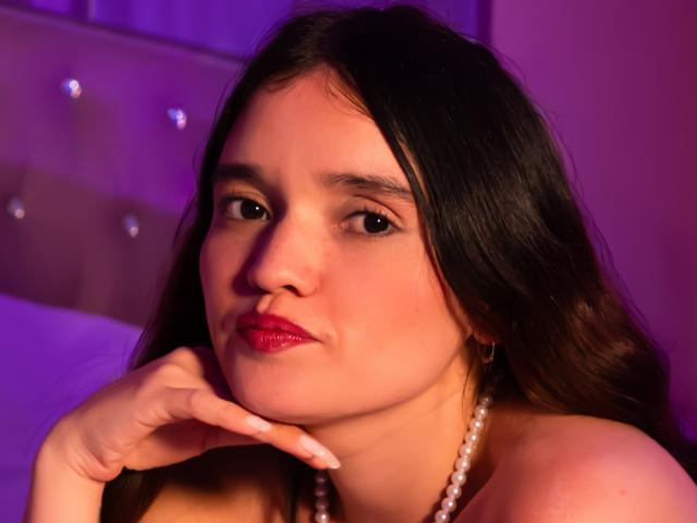AnabelleRoss with dark hair, red lipstick, pearl necklace, and hand on chin, looking at camera in purple light, showing her soft, curvy face and subtle makeup.