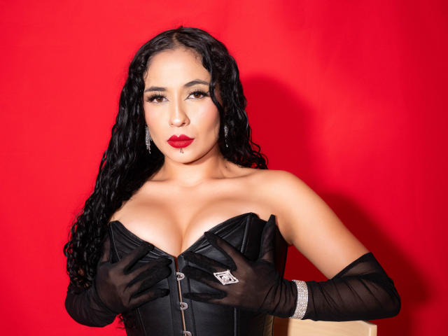 Lilith Stygian, con cabello negro y rizado, vestida con un corsé de cuero negro sin tirantes, guantes negros y lápiz labial rojo, cubriendo sus grandes senos con las manos, sobre un fondo rojo..