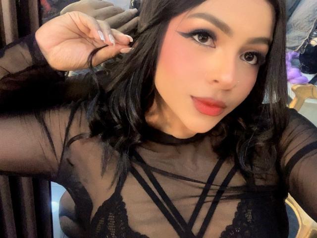 LindaFrancoo, avec un haut en dentelle noire, des manches transparentes et un rouge à lèvres rose, mettant en valeur sa poitrine, ses cheveux foncés et son eye-liner audacieux, tout en se tenant les cheveux. Léger sourire, adorable..