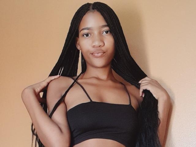 Plumm, con largas trenzas negras, sonriendo, vestida con un top negro sin tirantes, sosteniendo su cabello, mostrando sus pequeños senos y su piel morena, frente a una pared beige..