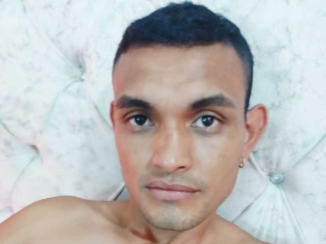 AndreyJurado tiene un rostro de tono medio, sin camisa, con cabello negro corto, pequeños aretes y una expresión neutra, mirando directamente a la cámara..