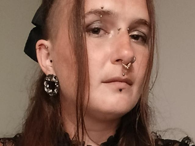 NightmareRaven hat mehrere Piercings im Gesicht: Nase, Nasenflügel und Oberlippe. Sie trägt ein schwarzes Spitzenoberteil und einen schwarzen Ohrring mit einem Stein, sowie eine schwarze Haarschleife..