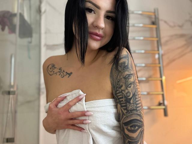 AuraXo, met zwart haar, houdt een witte handdoek voor haar borsten, heeft een tatoeage op haar linker schouder en arm, en een neusring, en ze glimlacht zachtjes..