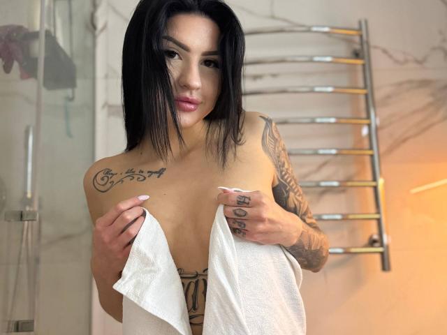 AuraXo, con cabello negro, tatuajes en los hombros y brazos, sosteniendo una toalla blanca que cubre sus senos, de pie en un baño con un tendedero de metal en el fondo..