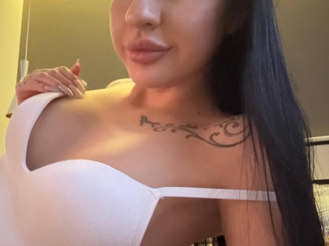 AuraXo, con i suoi lunghi capelli neri, indossa un top bianco senza spalline che mette in mostra il seno. Ha un tatuaggio sul petto ed è sorridente, con una mano vicino alla spalla..