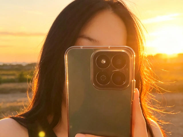 ValeriaRossii with long dark hair, holding a green iPhone in front of her face, sun setting in the background, golden light, visible eyes partially covered by the phone.