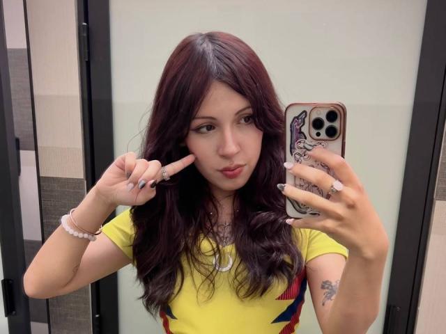 ValeryBeaumont with dark red hair, yellow top, and tattoos, making a peace sign with her right hand, holding a phone with a purple character case, in a mirror selfie.