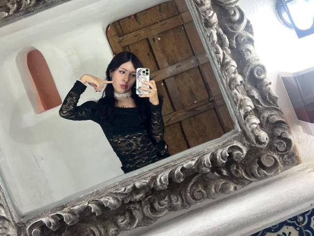 ValeryBeaumont with long black hair, wearing a black lace dress, pouting, pointing at lips in mirror selfie, holding phone with white case, ornate silver mirror frame.