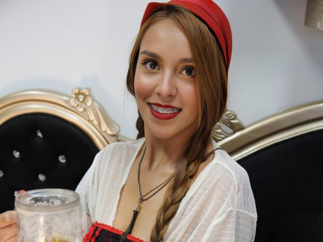 NaughtyAnny con i capelli castani raccolti in una treccia, un copricapo rosso, una maglia bianca trasparente, biancheria intima rossa e nera, che tiene un bicchiere, sorride con il rossetto rosso, mostrando l'apparecchio ortodontico..