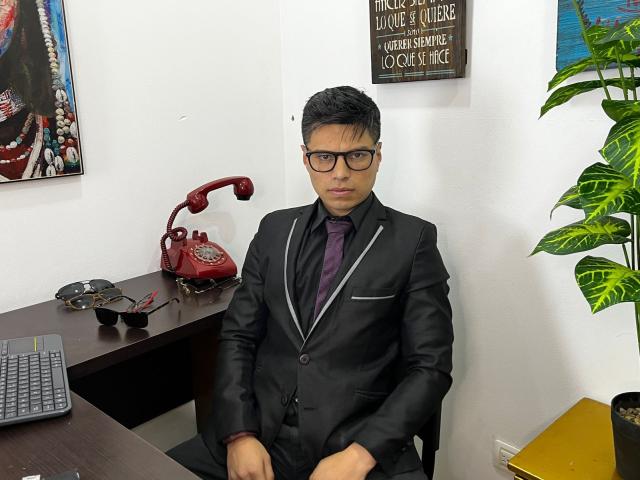 AquilesTaylor, wearing a black suit with purple tie, black glasses, short black hair, standing in front of a desk with a red phone, keyboard, and plant.