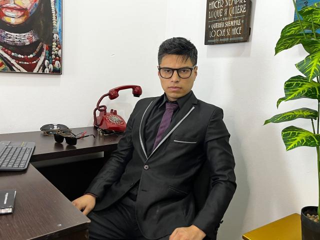 AquilesTaylor in a black suit with a purple tie, sitting at a desk, wearing glasses, with a red phone and a green plant in the background.