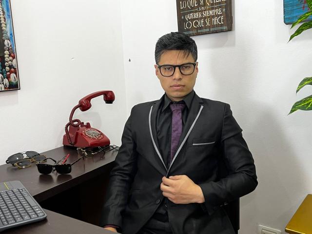 AquilesTaylor with short black hair, glasses, wearing a black suit, purple tie, and black shirt, standing in an office, holding his suit jacket, red phone, and sunglasses on desk.
