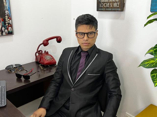 AquilesTaylor in a black suit with purple tie, sitting at a desk, wearing black glasses, red phone, glasses, and a black shirt, looking serious.