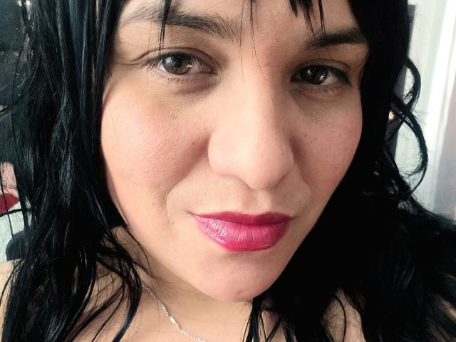 VivianaThompson has black wavy hair, brown eyes, and pink lipstick. Her fair skin is slightly textured with a small nose and subtle smile. She's wearing a silver necklace.