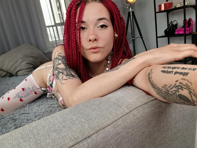 RachelNervis with red braids, white thigh-highs with red hearts, tattoos on arms, lying on gray sofa, pouty expression, crossed arms, wearing floral lingerie, in a cozy room.