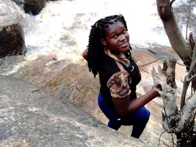 Mulher de pele escura, com cabelos longos trançados, sorrindo, vestindo uma camisa preta, uma manga camuflada, calças azuis, ajoelhada perto de uma rocha, segurando um galho de árvore, com uma cachoeira branca ao fundo..