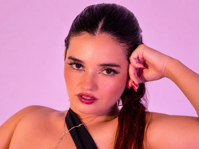 AnabelleRoss has her brown hair tied back, wearing a black halter top, with red nails and a silver chain necklace, looking at the camera with pink lipstick.