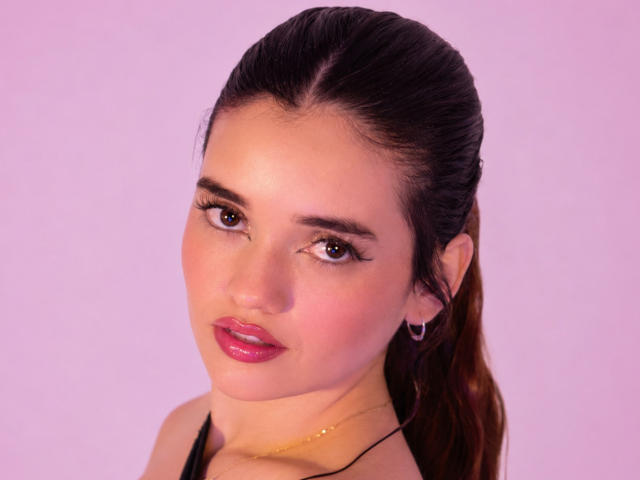 AnabelleRoss has pink lipstick, brown eyes, and a slight smile with glossy skin. She's wearing a black top and a gold necklace, with her brown hair in a ponytail.
