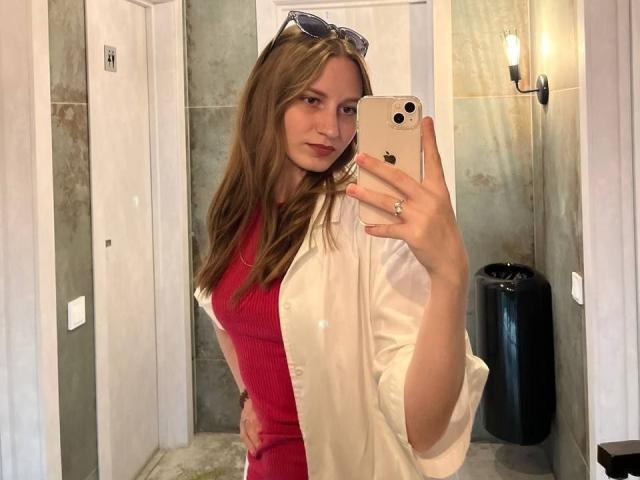 Barbara Hearns, com cabelos castanhos longos, vestindo uma blusa vermelha e uma camisa branca, segurando um iPhone, tirando uma selfie no espelho, com óculos de sol na cabeça, em um banheiro..