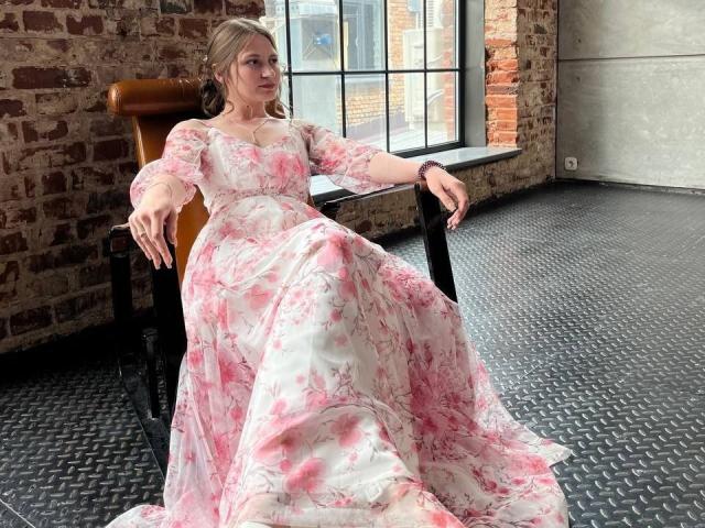 Barbara Hearns, vestita con un abito floreale rosa, è seduta su una sedia marrone con le gambe incrociate, le braccia appoggiate sulla sedia, in una stanza con una parete in mattoni..