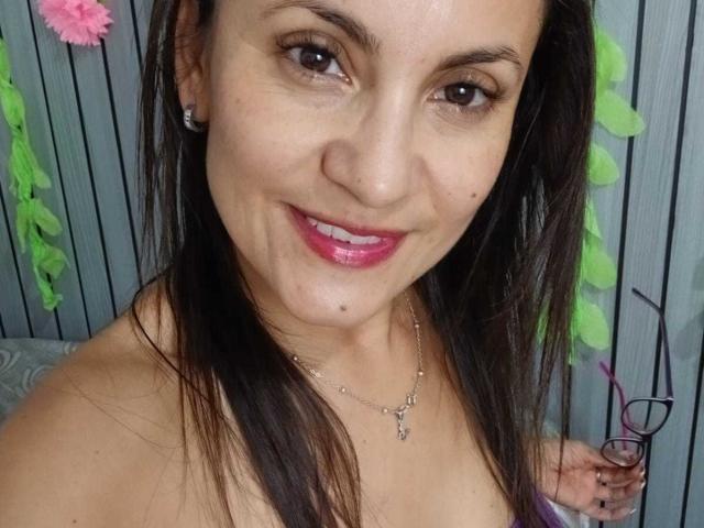 SolaraDubois with pink lipstick, brown eyes, and straight dark hair, smiling, showing small mole under her chin, wearing a necklace, holding glasses, and a pink flower in the background.
