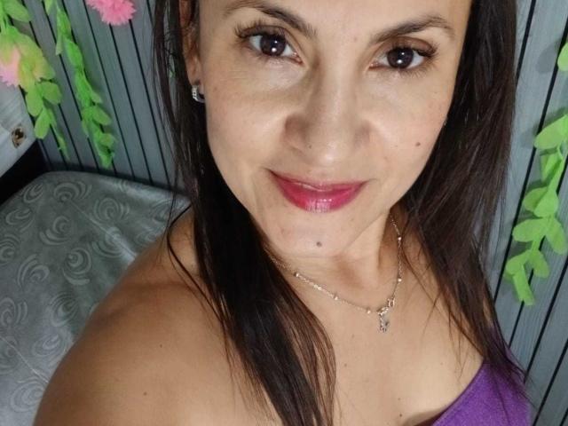 SolaraDubois with pink lipstick, brown eyes, and long dark hair, wearing a purple top, showing off her tits and a small mole under her right eye.