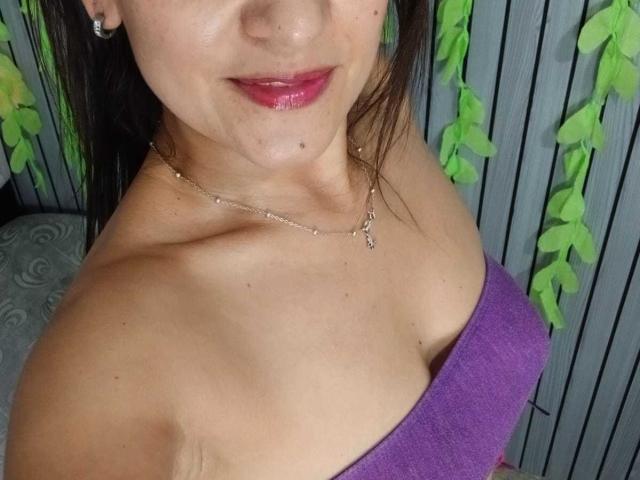 SolaraDubois with shiny pink lipstick, slight smile, bare shoulder, purple strap dress, silver necklace, visible collarbone, and small mole on her cheek. Her tits are partially visible.