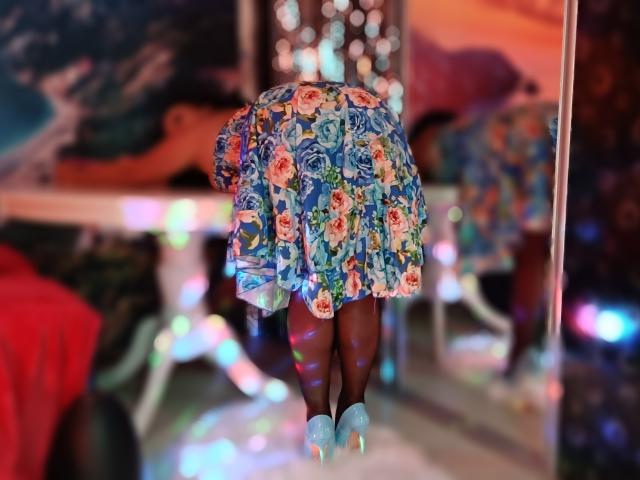 AmberSunshine stands with her back to the camera, wearing a colorful floral dress and blue high heels, her dark skin visible, reflecting in a mirror with bokeh lights.