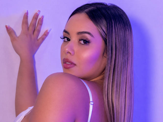 ZoeVibe with long blonde hair, looking over her shoulder, wearing a white bra, hand against a wall, purple-blue lighting, glossy pink lips, and manicured nails.