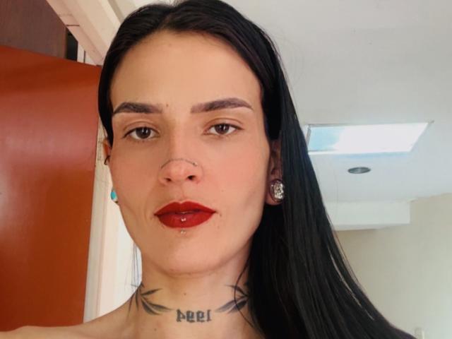 EmmaHerby has long black hair, red lipstick, a nose ring, and a tattoo under her chin with crossed lines. She's wearing ear studs and has a confident expression.