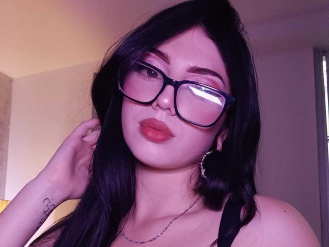 IsabellaaRossi with black hair, glasses, and red lipstick, touching her ear, wearing a black tank top, showing a small tattoo on her wrist.