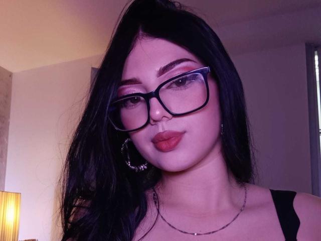 IsabellaaRossi with black hair, glasses, red lipstick, hoop earrings, and a black tank top, showing her pouty ass-kissing face in a purple-lit room.
