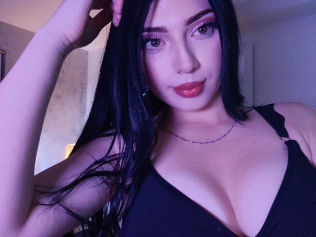 IsabellaaRossi with black hair, blue eyes, and red lipstick, wearing a black bra, exposing her large tits, hand in hair, slight smile, wearing a thin necklace.