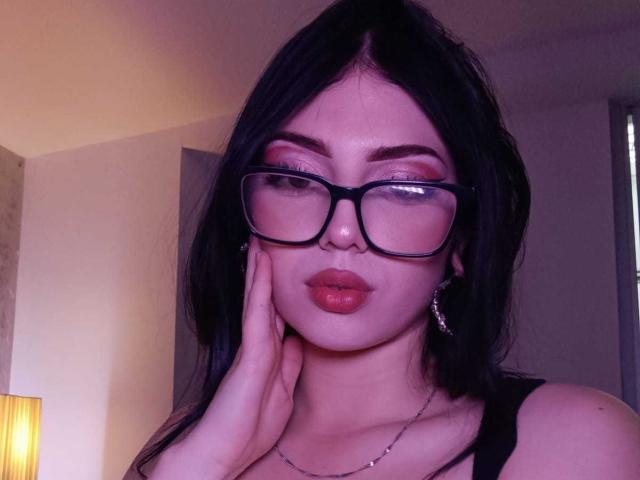 IsabellaaRossi with black hair, glasses, and red lipstick, hand on cheek, wearing a black top, small silver necklace, and a thin chain earring, in a softly lit room.
