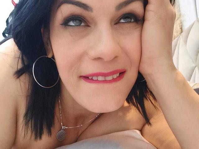 Liza Russell tem cabelo preto, olhos azuis e batom rosa, com a cabeça apoiada na mão, sorrindo, com os mamilos rosa quase imperceptíveis, usando brincos grandes e um colar..