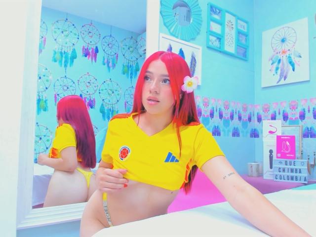 MiaLeroy with red hair, yellow crop top, and yellow thong, showing her small tits and bare ass, holding a yellow marker, reflected in a mirror, in a bright room.