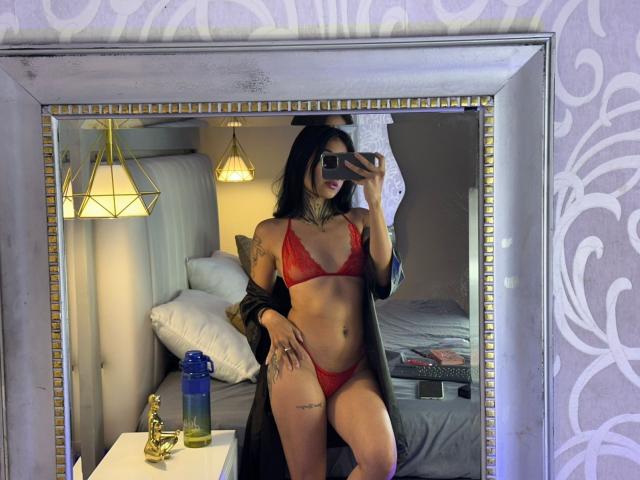 NicolleSweetty vestindo um sutiã e calcinha vermelhos de renda, mostrando seus seios e vulva, com cabelos pretos longos, tatuagens e um robe preto, tirando uma selfie no espelho..