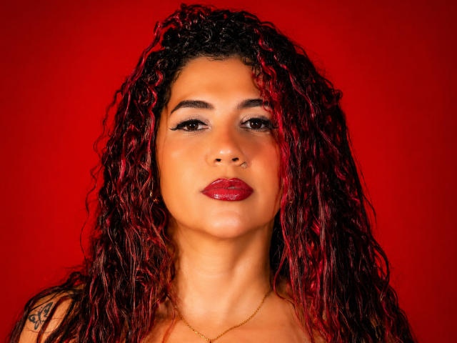 Antonela Riveiro tem cabelos ruivo-rosados e cacheados, sobrancelhas escuras, batom vermelho e um piercing no nariz. Ela está usando um colar dourado contra um fundo vermelho sólido..