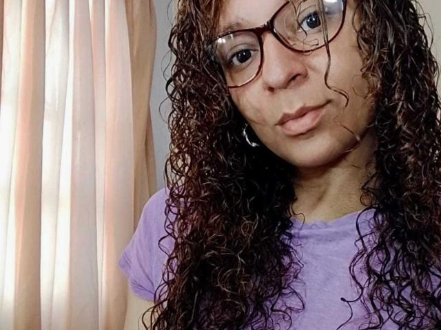 Angelonix with curly brown hair, wearing purple shirt and glasses, pouting slightly, showing small tits, standing near white curtains.  She's got a cute face and a slight smile.