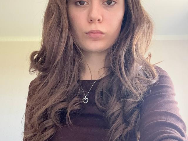 Arlene Giannone, con cabello castaño largo y ondulado, vistiendo una blusa marrón y un collar de plata con forma de corazón, con una expresión seria y los labios ligeramente entreabiertos, sin maquillaje, en un selfie..