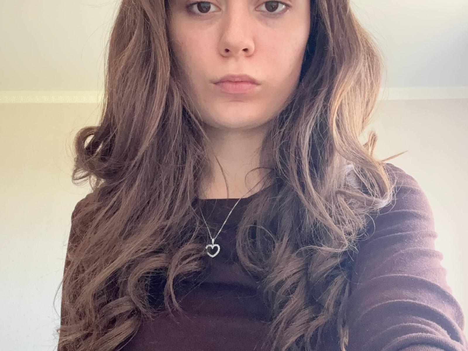 ArleneGiannone with long brown wavy hair, wearing a brown top and a silver heart necklace, looking serious with slightly parted lips, no makeup, in a selfie.