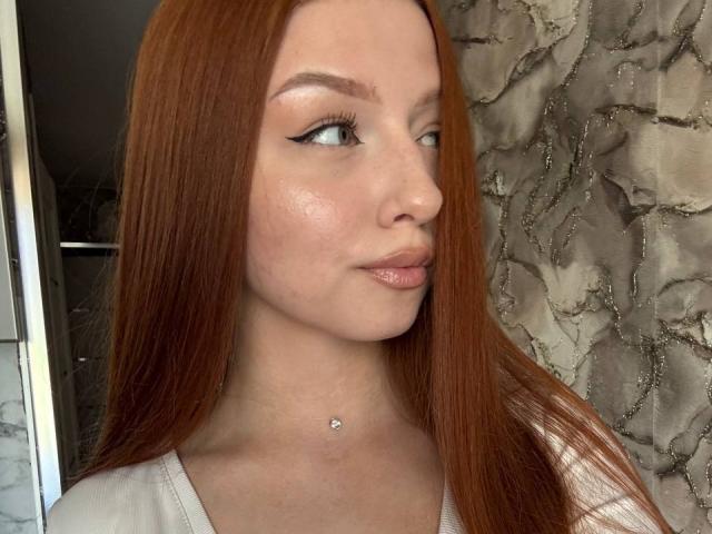 LuanneKadri with red hair, white eyeliner, shiny skin, small nose piercing, glossy pink lips, wearing a white top, looking to the side, in a selfie.