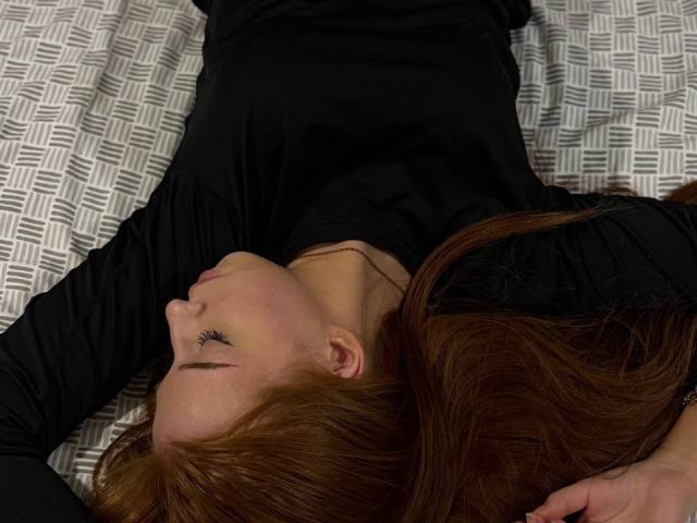 LuanneKadri è sdraiata sulla schiena con i lunghi capelli rossi sparsi, indossa una maglia nera a maniche lunghe, ha gli occhi chiusi e porta una sottile collana. Il suo viso è leggermente arrossato..