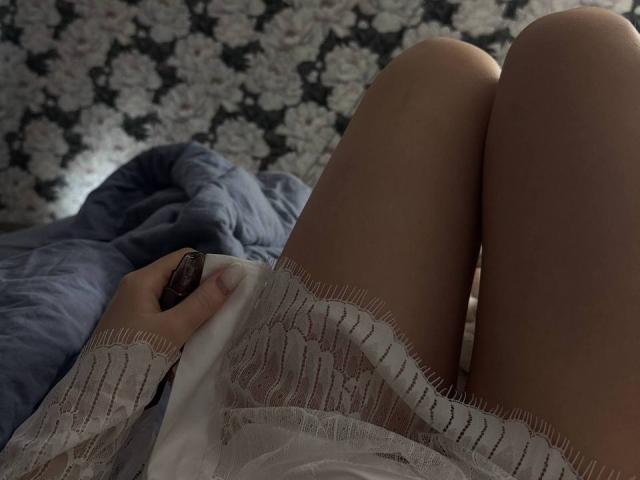 LuanneKadri staat met gespreide benen, gekleed in een witte kantjurk, en houdt een afstandsbediening vast. Aan de linkerkant ligt een blauwe deken en op de achtergrond is bloemenbehang te zien..