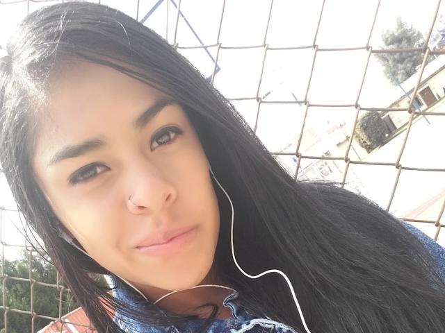 StellaBeckker a de longs cheveux noirs, un anneau de nez et des écouteurs, avec un léger sourire, elle porte un sweat à capuche bleu, sous une lumière du soleil vive, et regarde l'appareil photo..