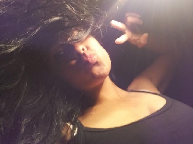 StellaBeckker with dark, wavy hair covering her eyes, pouting her lips, wearing a black tank top, making a peace sign with her right hand in a dimly lit photo.
