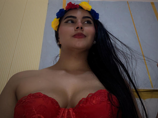 AlaraKey with long black hair, wearing a red lace bustier, red lipstick, and a yellow-blue flower headband, looking up with a slight smile. Boobs visible, medium-sized.