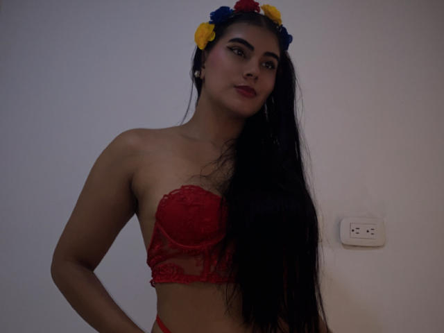 Alara, con cabello negro largo, un sujetador de encaje rojo y una corona de flores, mirando a la cámara, con las manos en las caderas, mostrando sus pechos, de pie frente a una pared blanca..