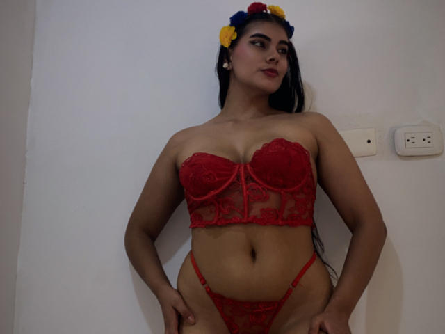 AlaraKey, con cabello negro largo, vistiendo un sujetador y tanga de encaje rojo, una corona de flores, de pie, con las manos en las caderas, mirando hacia un lado, lápiz labial rojo, busto mediano, piel morena..