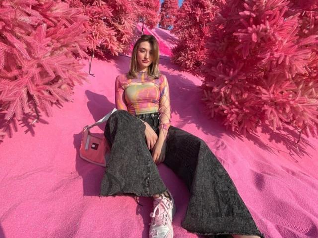 Kevin Stroll mit rosa Bäumen, der ein farbenfrohes Tie-Dye-Oberteil, schwarze, weite Hosen und weiße Turnschuhe trägt, sitzt auf rosa Boden, hält eine rosa Tasche und hat einen entspannten, sexy Look..